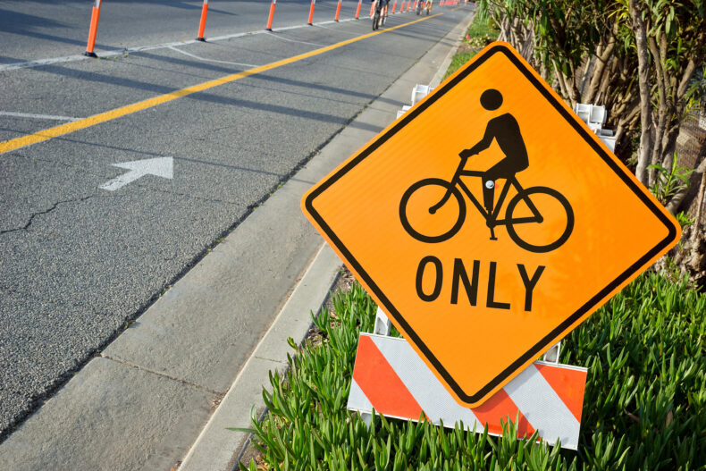 Side of the road has bicycle lane mark and cone off to oncoming cars. This Bicycle sign is also on side of the road for communication awareness and traffic management